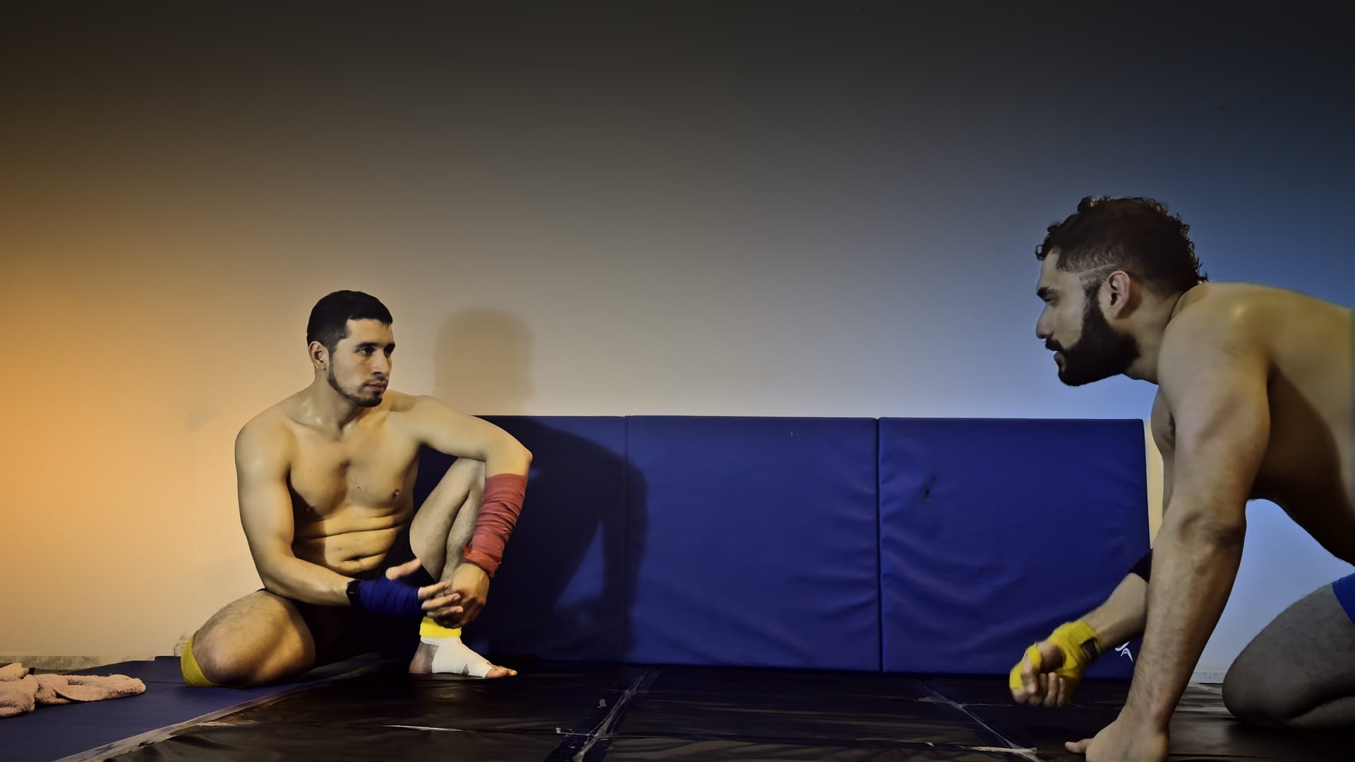Nocturmex VI face-off before the match: Nocturmex seated against the wall pads, Robgrappler on his knees across the mat