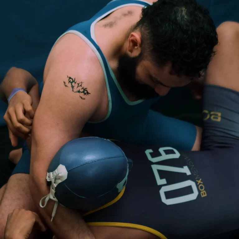 RobGrappler in a submission grappling match, wearing a blue singlet with a tattoo, against an opponent in a blue suit with the number 076, on a mat with a blue backdrop.